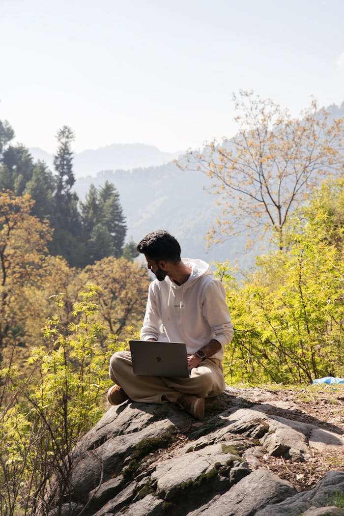 A man in a hoodie sits on a rock with a laptop, surrounded by forested mountains.